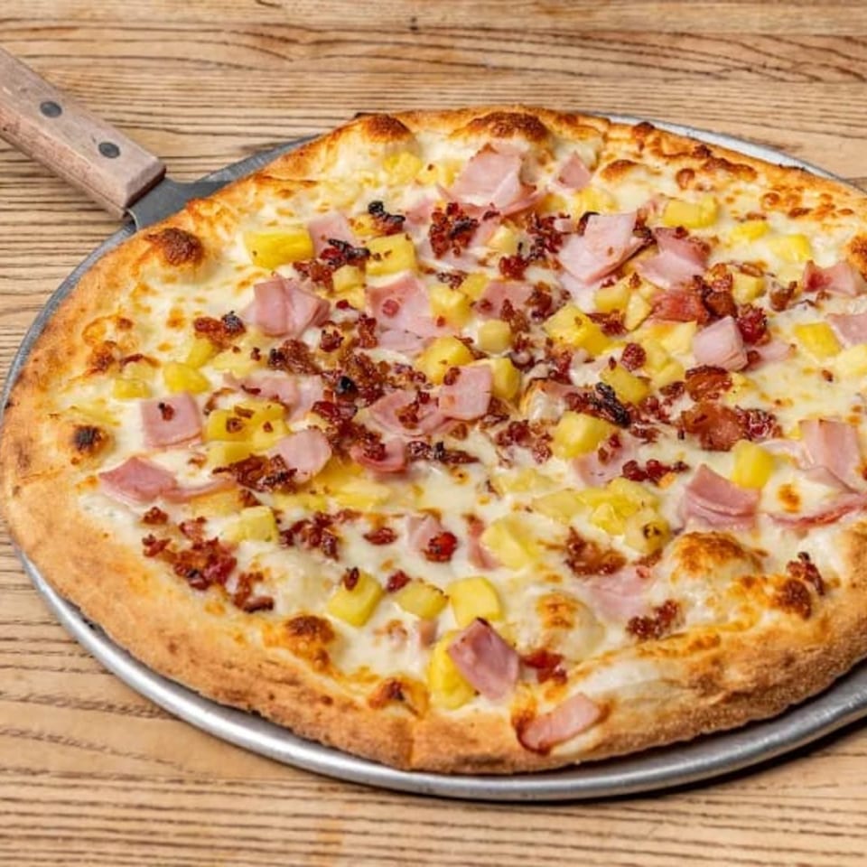 Hawaiian pizza with ham, pineapple, and bacon on a metal pizza peel, on a wooden table.